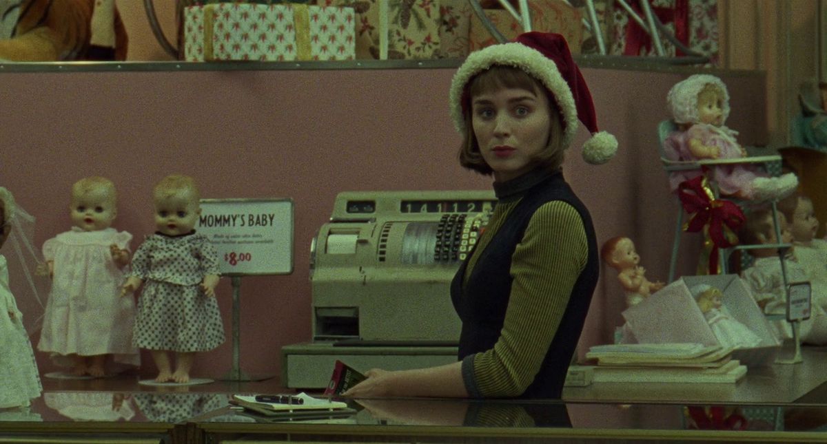 Rooney Mara indossa un cappello da Babbo Natale dietro il bancone di un negozio.  Sullo sfondo sono sparse delle bambole e un cartello recita 