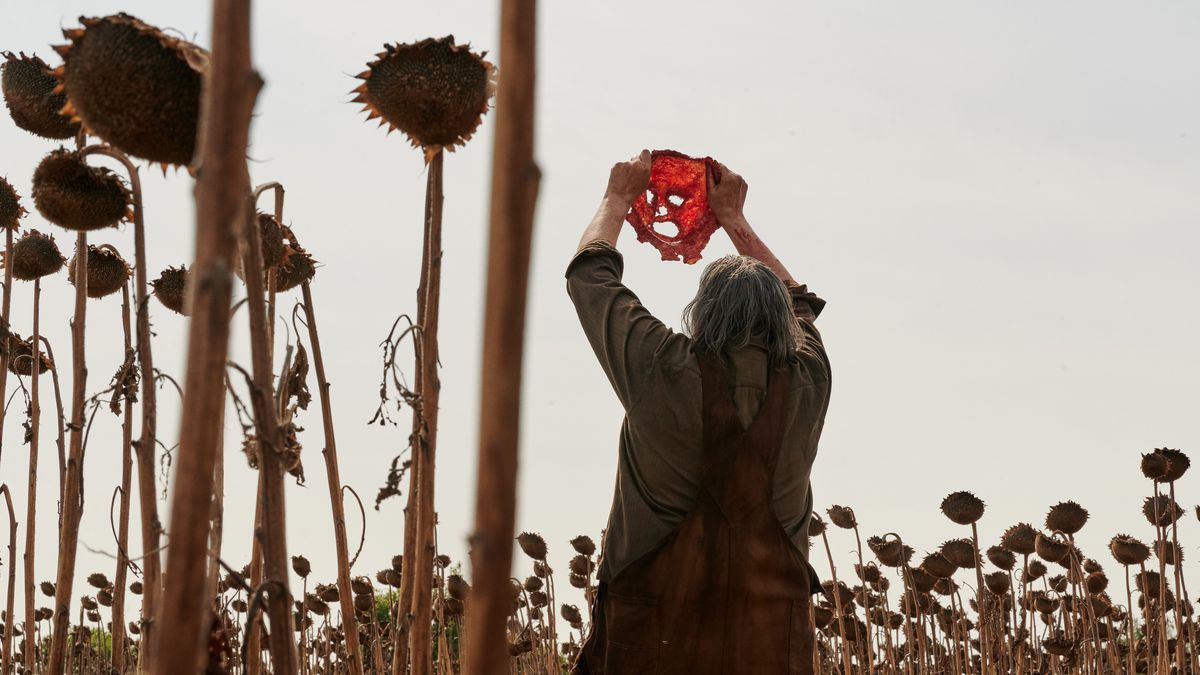 Leatherface (Mark Burnham) è in piedi con le spalle alla telecamera in un campo di girasoli morti e tiene in alto la sua maschera di pelle in modo che la luce del sole brilli attraverso gli occhi, il naso e la bocca nel massacro del Texas Chainsaw del 2022