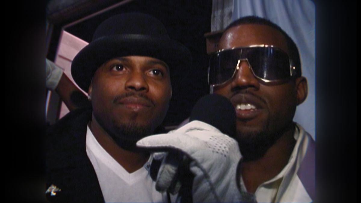 un'immagine dal primo episodio di jeen-yuhs: A Kanye Trilogy of Kanye West, indossa occhiali da sole e tiene un microfono con un guanto bianco, in piedi accanto a un altro uomo di colore che indossa un fedora