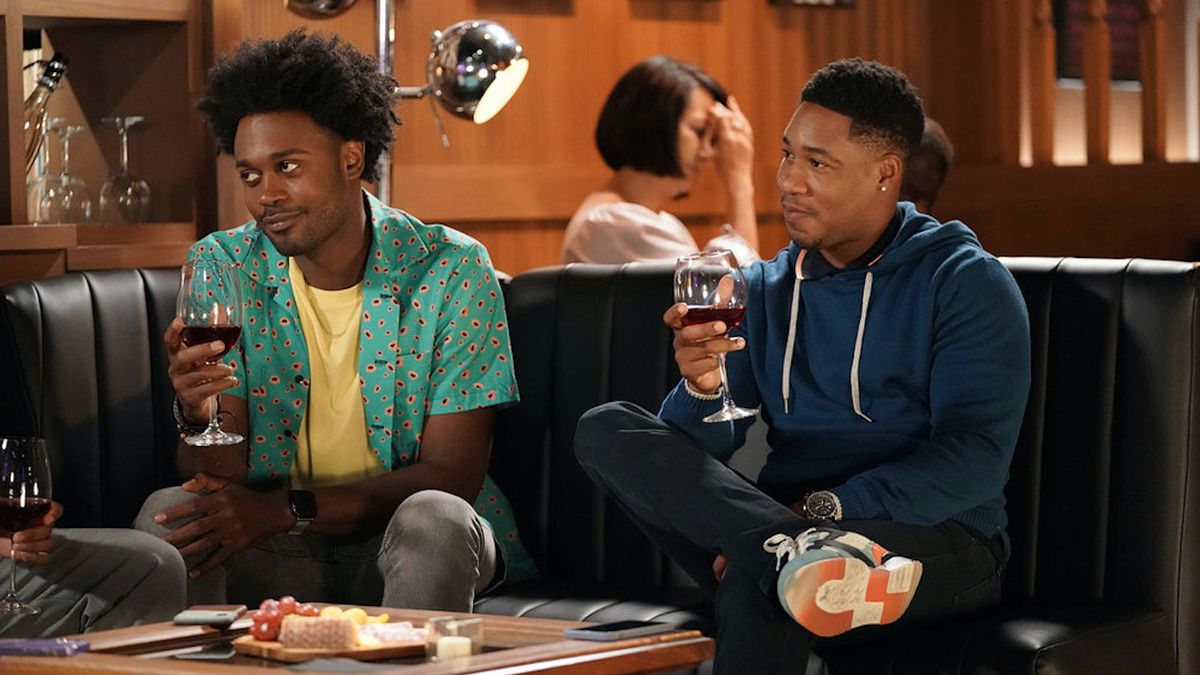 Due membri del cast dello spettacolo Grand Crew si siedono su un divano a bere vino con un tagliere di formaggi sul tavolo di fronte a loro.