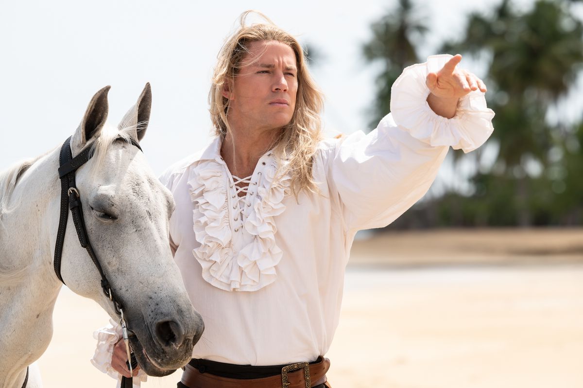 Channing Tatum, con una camicia bianca a balze, indica in lontananza mentre è in piedi con un cavallo bianco su una spiaggia nella città perduta