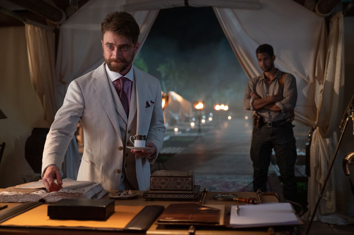 Daniel Radcliffe in abito bianco tiene una tazza di tè e sta in piedi sopra una scrivania in una tenda nella Città Perduta