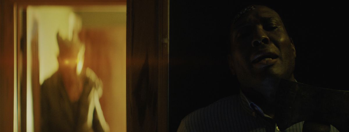 Geno Walker nei panni di Ken Barber che impugna un'ascia arrugginita e si nasconde da un demone con occhi luminosi in Night's End.