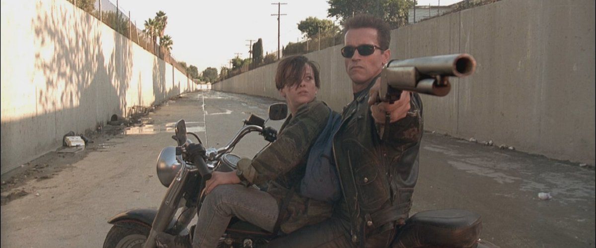 (LR) Edward Furlong e Arnold Schwarzenegger nei panni di John Conner e un Terminator T-800 riprogrammato che impugna un fucile in cima a una motocicletta ferma in un burrone in Terminator 2: Judgment Day. 