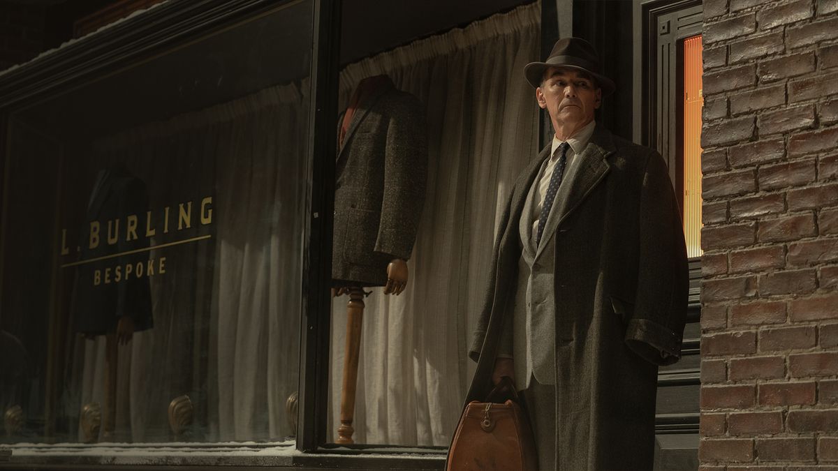 Mark Rylance nei panni di Leonard in abito e cappotto in piedi fuori dalla sua sartoria all'angolo in The Outfit (2022).
