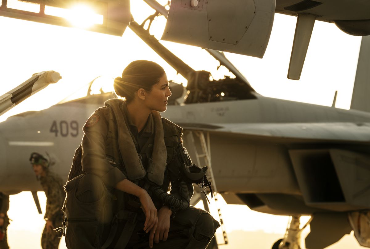 Monica Barbaro si accovaccia vicino ad alcuni jet da combattimento con il sole alle spalle