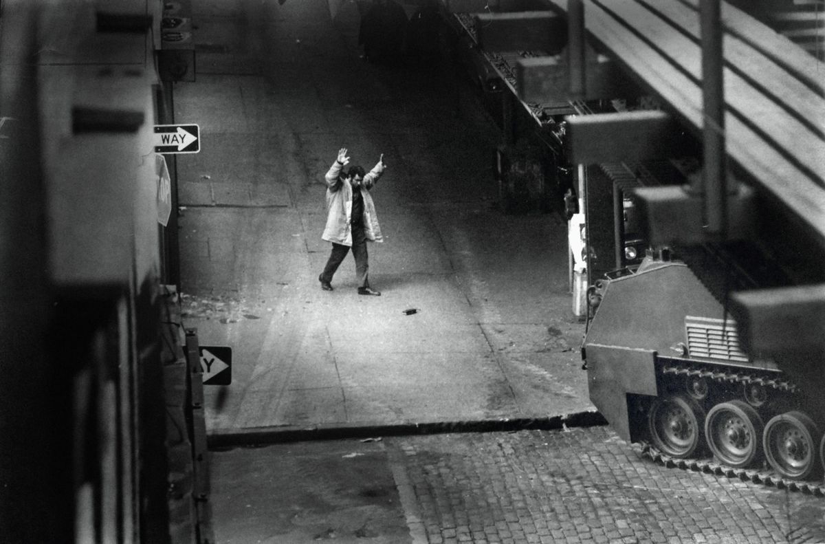 Un uomo con un trench si tiene le mani sopra la testa all'angolo di una strada in Hold Your Fire.