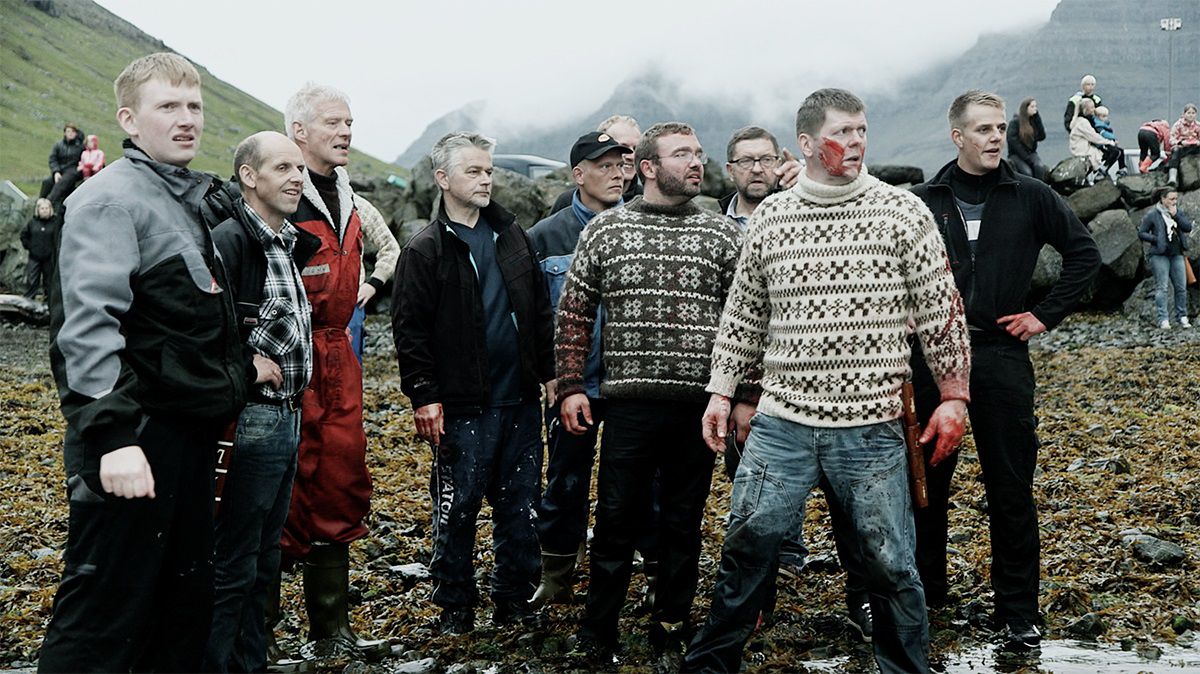 Un gruppo di uomini in felpa, uno dei quali coperto di sangue, si trova sulla riva di una spiaggia in A Taste of Whale.
