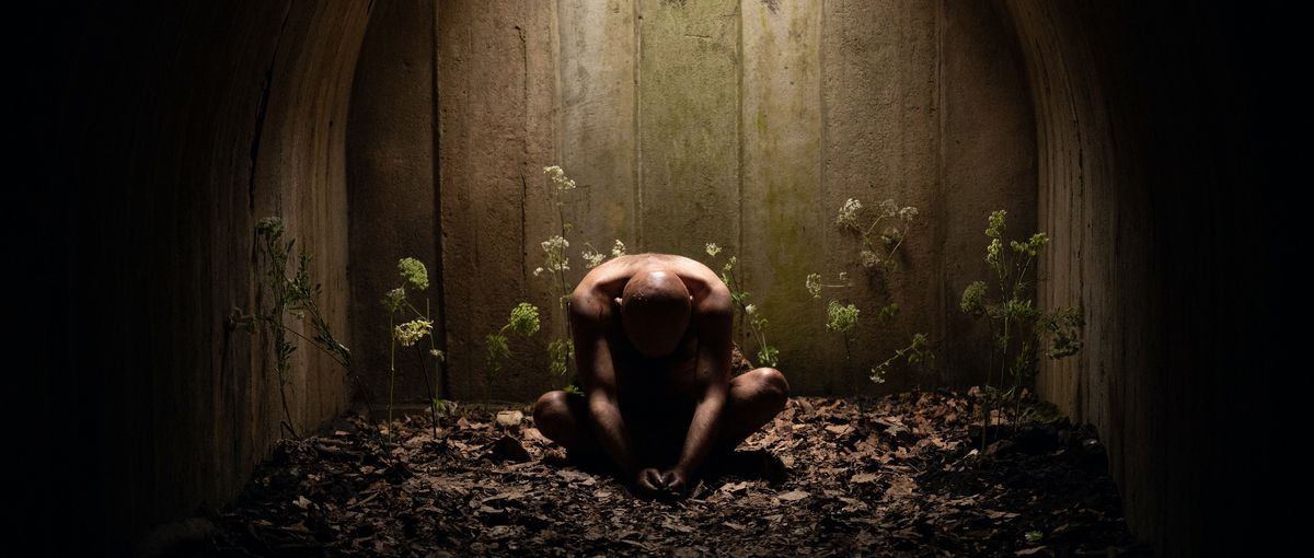 Rory Kinnear siede nudo, curvo, in un mucchio di foglie secche circondate da piante filanti in Men