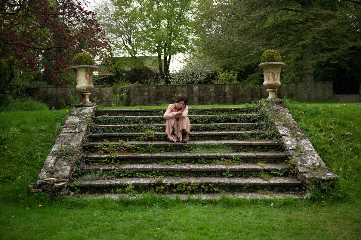 Jessie Buckley, con un lungo vestito rosa, siede su scale coperte di muschio in un paesaggio verde in Men di Alex Garland.