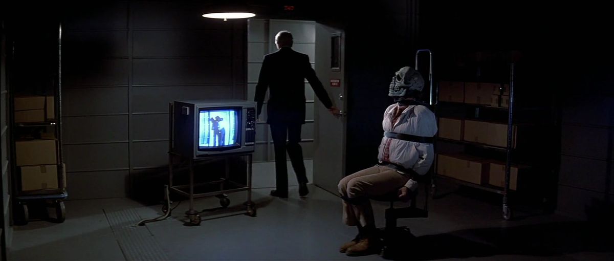 Un uomo legato a una sedia con una maschera da teschio è costretto a guardare la TV in Halloween III: La stagione della strega.
