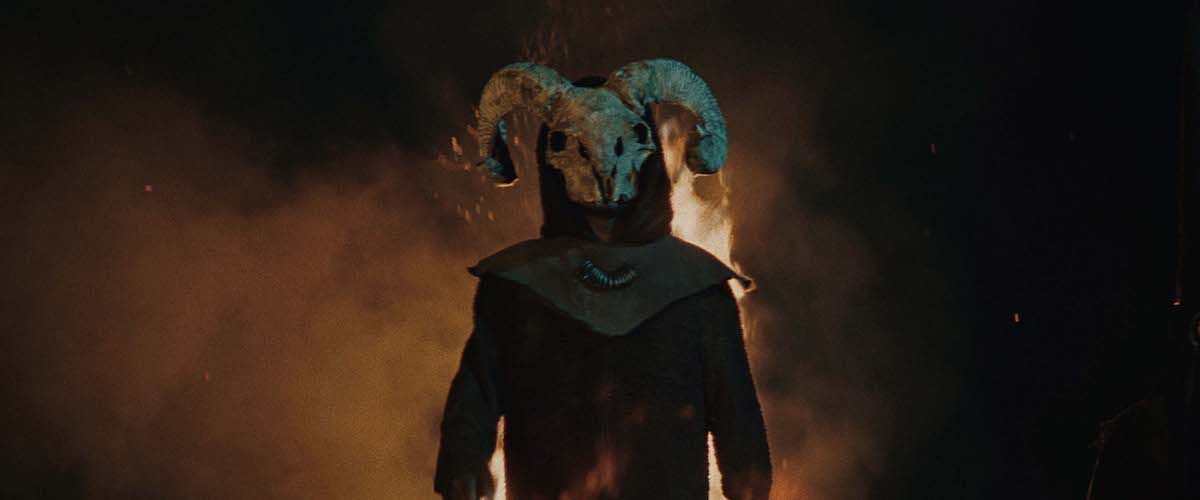 Una figura ammantata che indossa un teschio di ariete come maschera si trova di fronte a uno sfondo infuocato.