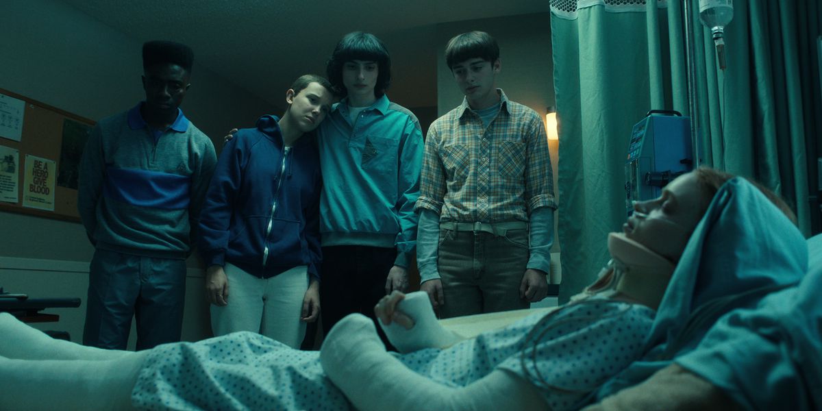 Lucas, Eleven, Mike e Will in piedi e guardando Max in un letto d'ospedale