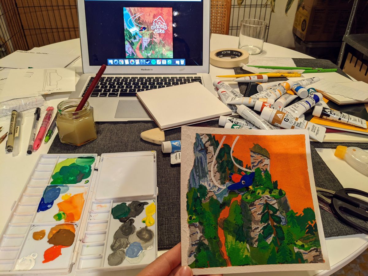 Un tavolo disordinato con un sacco di bottiglie di vernice sparse.  Sullo sfondo un laptop mostra un'immagine artistica per il gioco A Short Hike.  In primo piano, una tavolozza di colori contiene molti colori e accanto c'è un dipinto di una breve escursione.