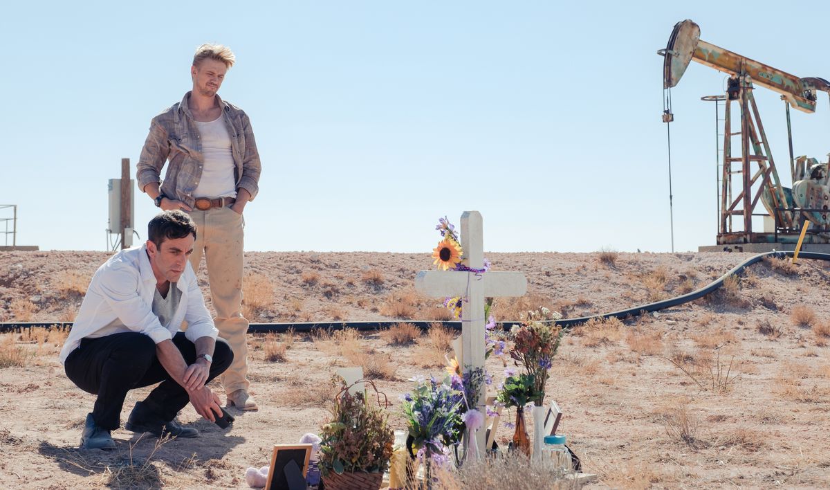 BJ Novak e Boyd Holbrook stanno vicino a una croce commemorativa disseminata di bouquet in un campo giallo sterile vicino a una torre di petrolio in Vengeance