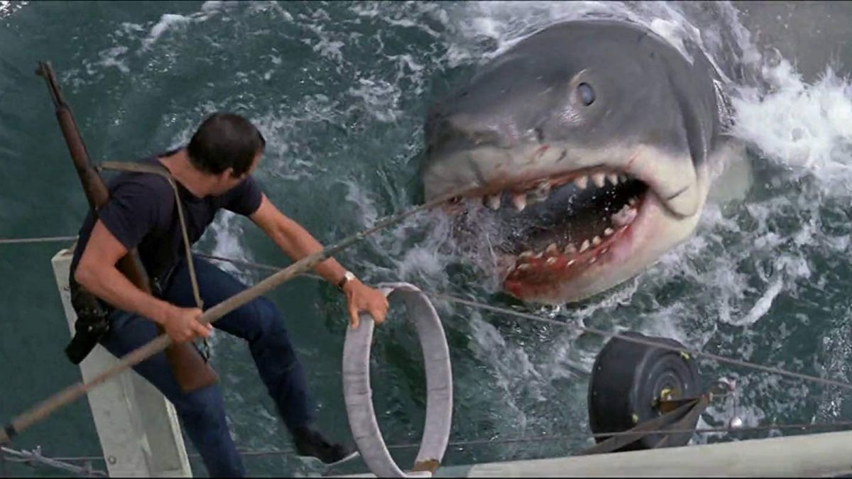 Un uomo con una lancia si trova sul lato di una barca che si ribalta in mare mentre fissa negli occhi il grande squalo bianco digrignando i denti in Jaws.