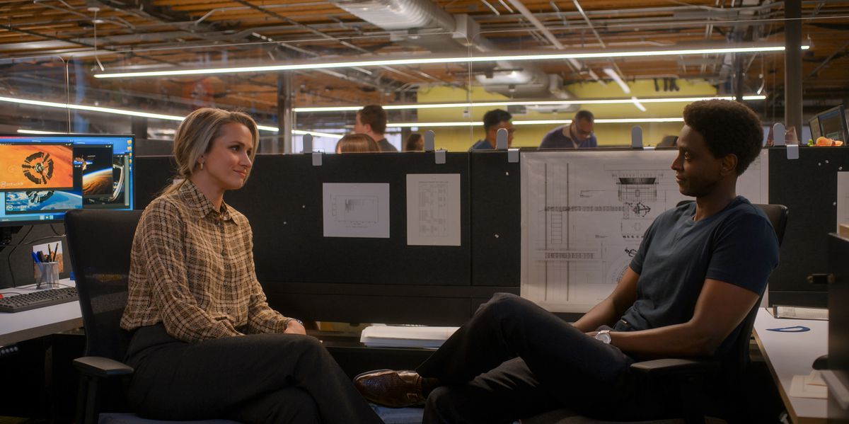 Karen e Dev seduti in un cubicolo dell'ufficio si sorridono