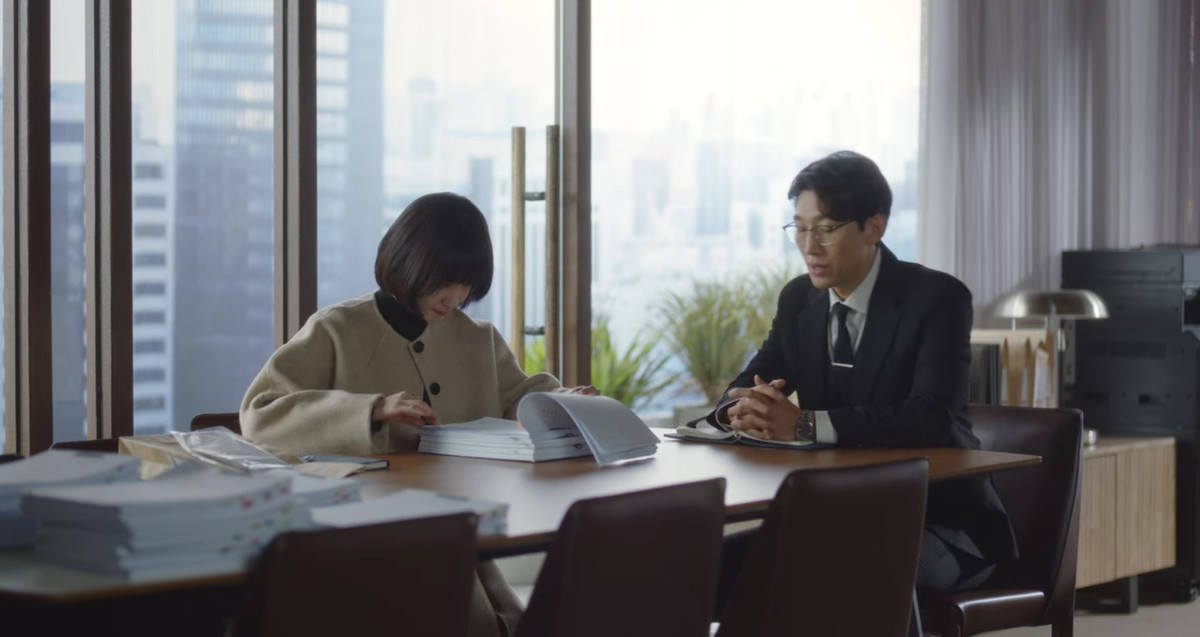 Woo Young-woo e il suo collega seduti a un tavolo da conferenza.  Sta studiando attentamente i documenti mentre lui è seduto e sembra leggermente costernato. 