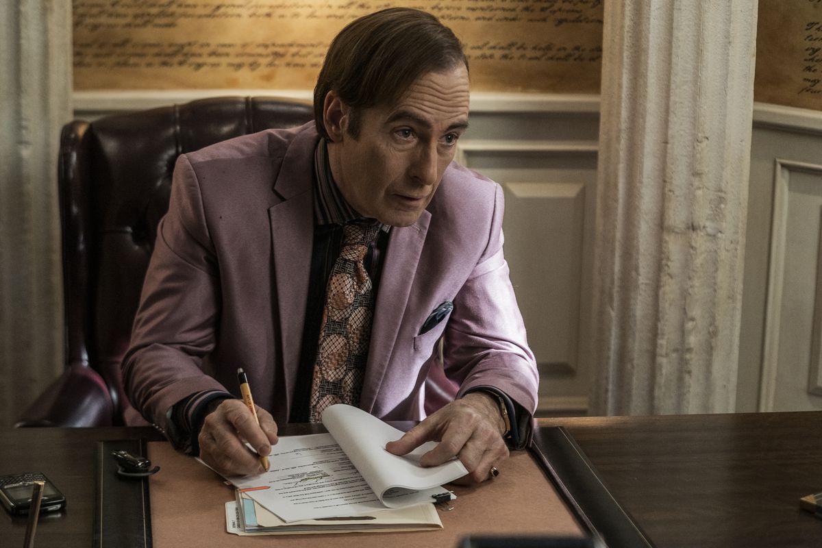 Saul Goodman seduto alla sua scrivania con un vestito viola che guarda qualcuno fuori dalla telecamera.  La telecamera è angolata verso di lui mentre firma i documenti per il divorzio.