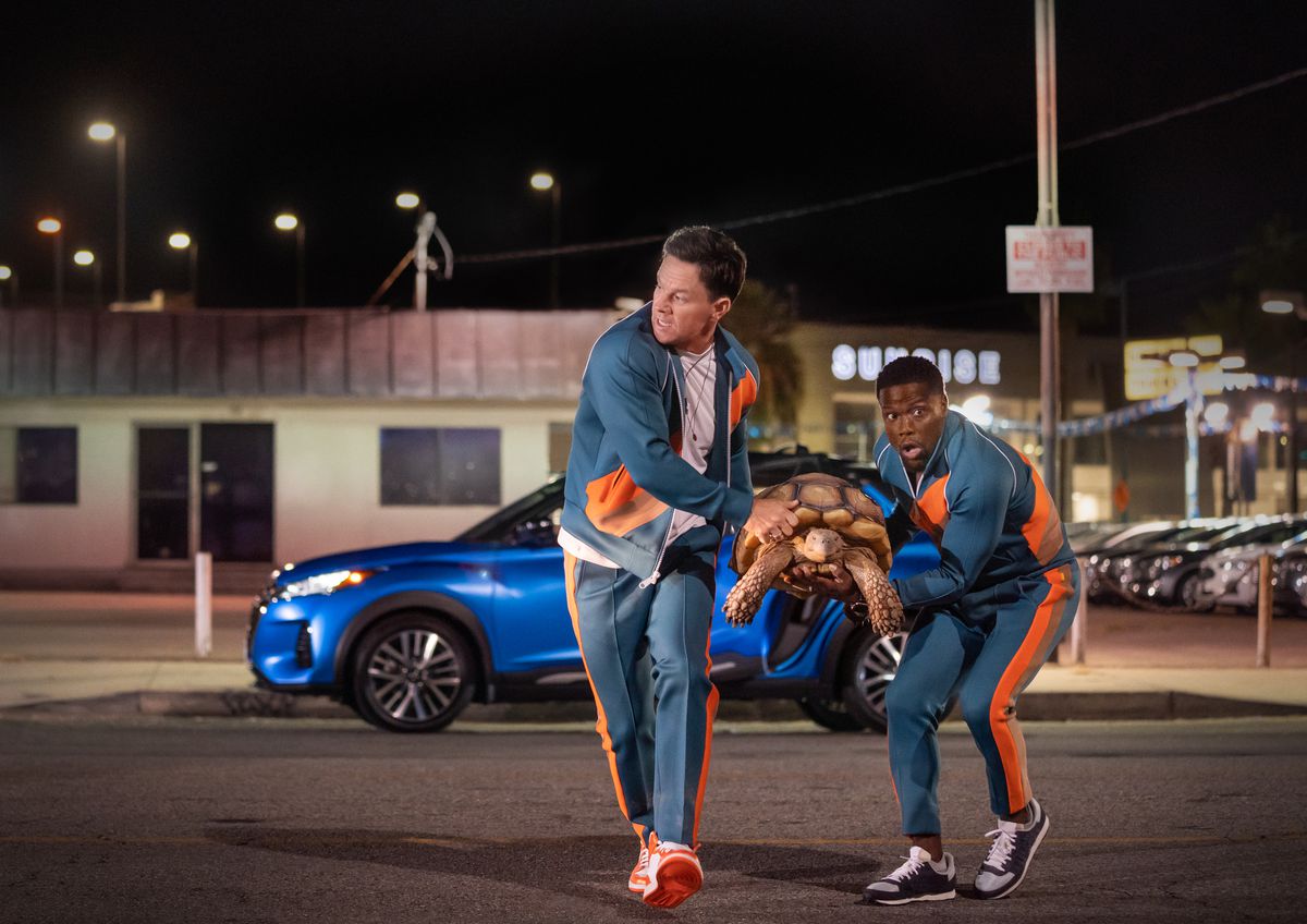 Mark Wahlberg e Kevin Hart nei panni di Huck e Sonny che trasportano una tartaruga da un minivan blu in tute blu e arancio abbinate in Me Time.