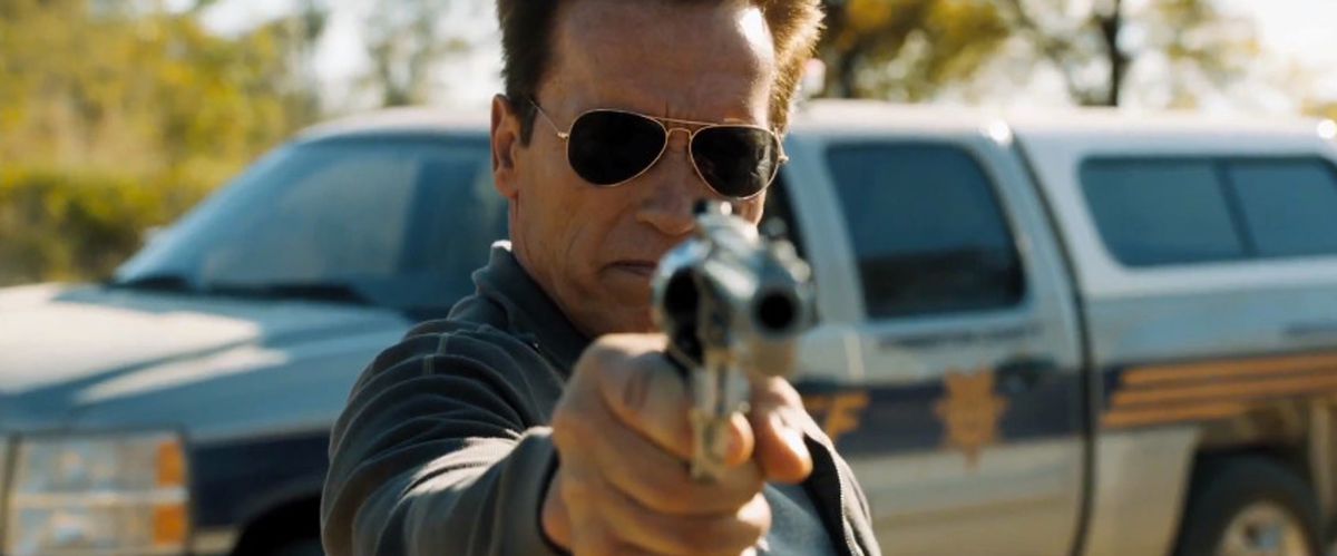Arnold Schwarzenegger, indossando occhiali da sole da aviatore e davanti al camion di uno sceriffo, punta un revolver verso la telecamera in The Last Stand.