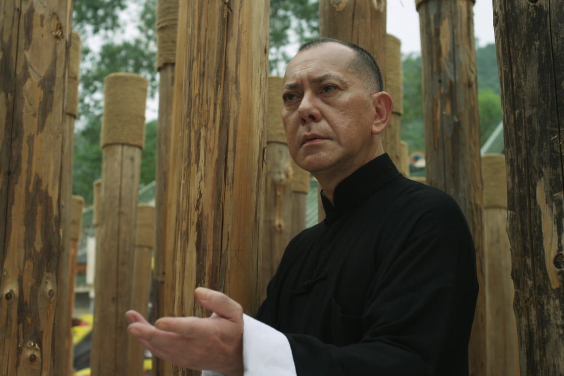 Anthony Wong indossa un tradizionale abito da kung fu nero con colletto e assume una posa difensiva davanti a bastoncini di legno in Ip Man: The Final Fight
