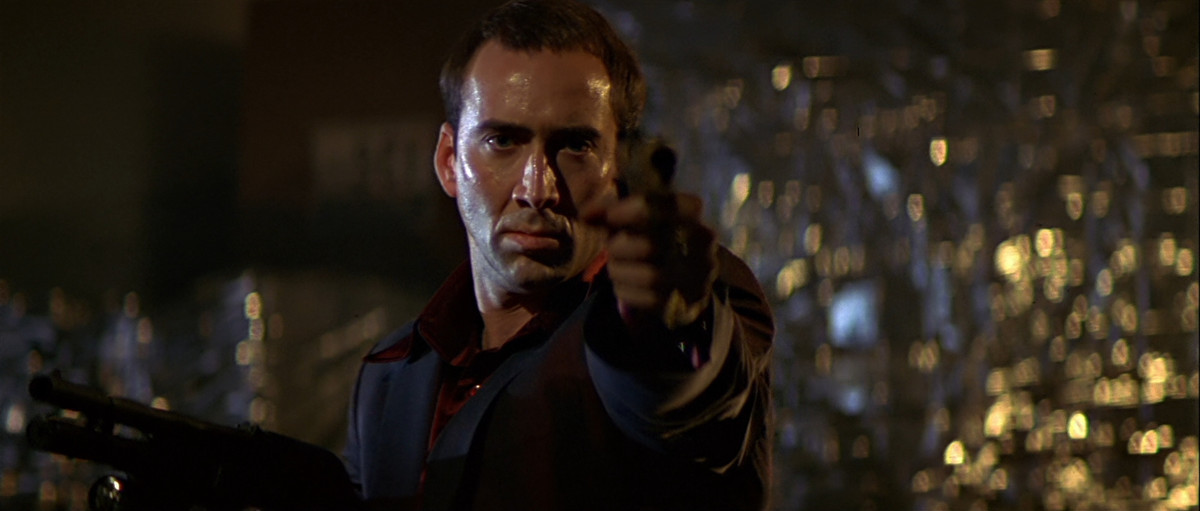 Un sudato Nicolas Cage in giacca e cravatta rossa punta una pistola verso la telecamera in Face/Off