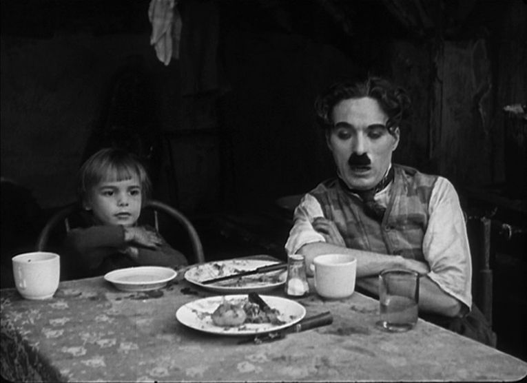 Charlie Chaplin e Jackie Coogan siedono con le braccia incrociate a un tavolo da pranzo in The Kid