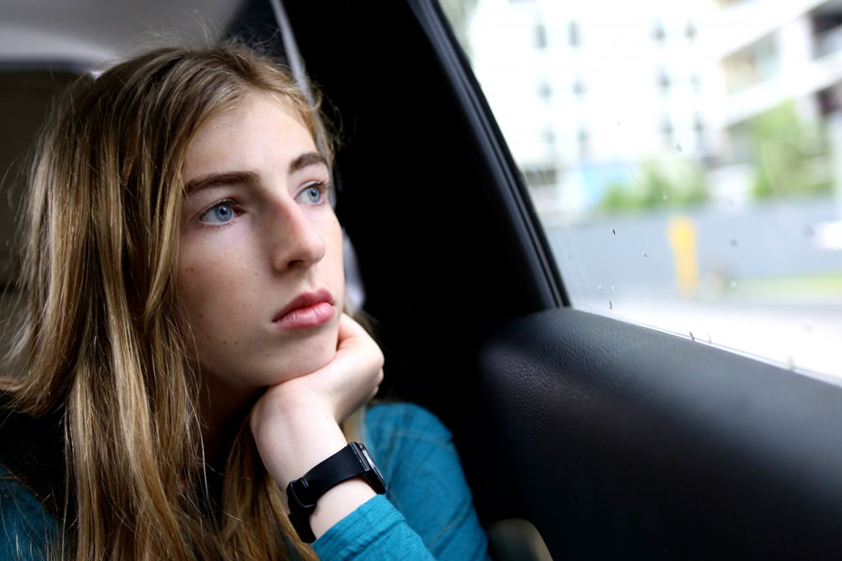 Una ragazza con i capelli biondi con una maglietta turchese a maniche lunghe che guarda fuori dal finestrino di un'auto.