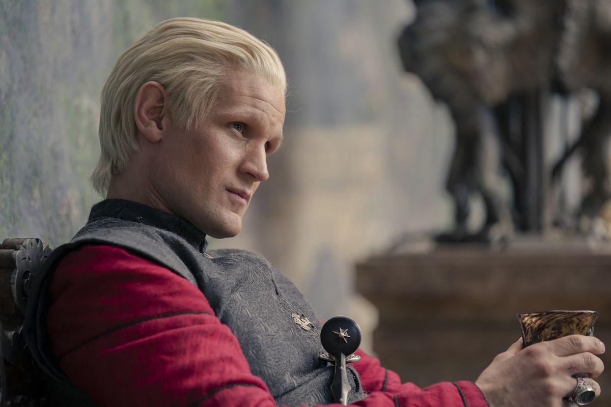 Daemon Targaryen, unto, con indosso una maglietta rossa e una tunica grigia, fissando qualcuno mentre stringe il suo vino in House of the Dragon