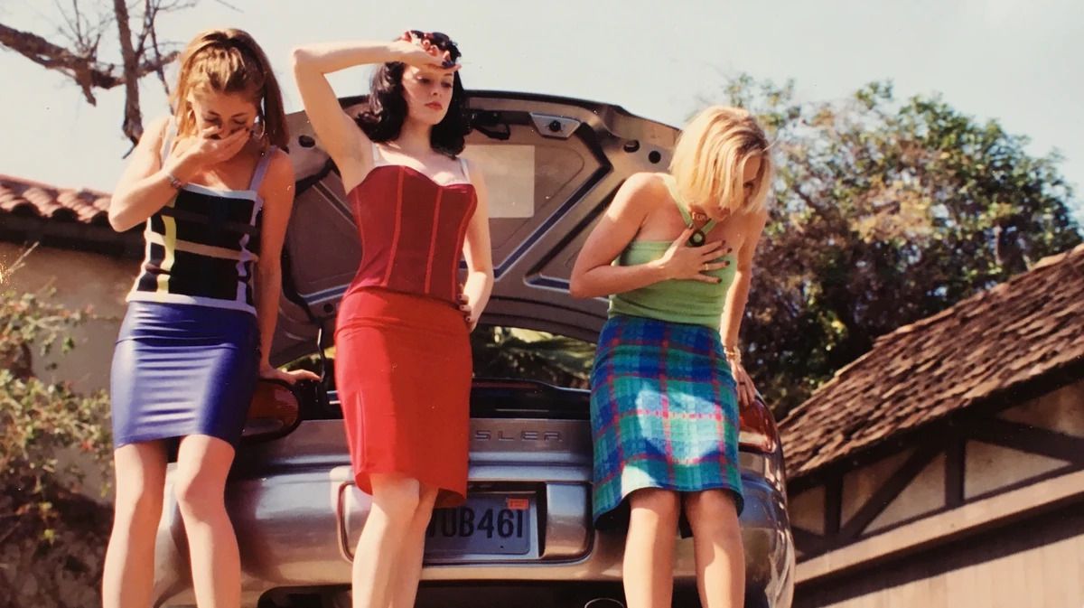 tre ragazze adolescenti dai colori vivaci stanno accanto al bagagliaio di un'auto aperto;  due di loro gag;  quello al centro, che veste di un rosso acceso, sembra composto e quasi annoiato