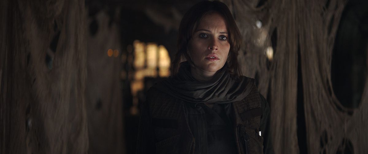 Jyn Erso, una donna dai capelli scuri che indossa una giacca e una sciarpa, emerge dall'ombra in Rogue One: A Star Wars Story
