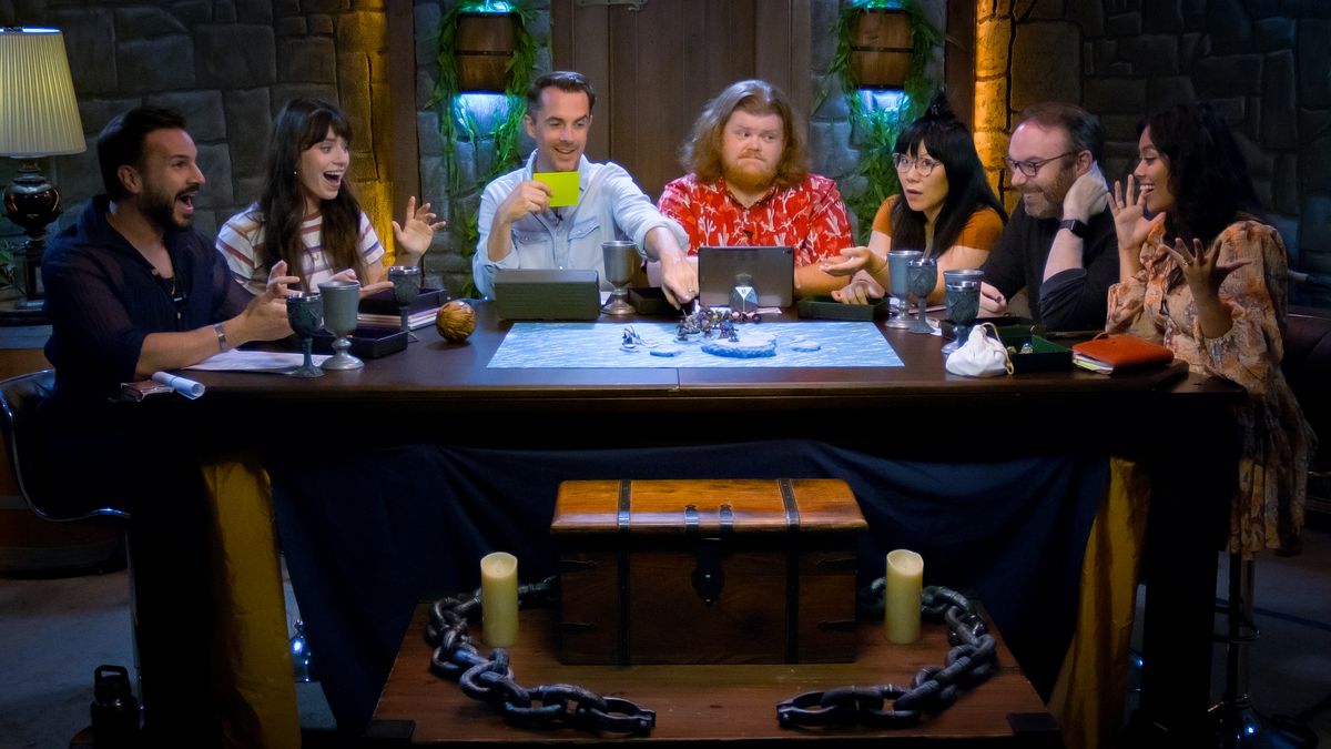 Sette membri del cast siedono attorno a un tavolo, tutti reagendo al gioco da tavolo in corso