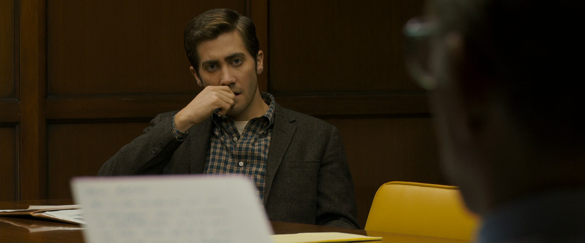 Jake Gyllenhaal, con indosso un abito e una camicia con colletto abbottonato, si morde le unghie mentre guarda un uomo che legge un pezzo di carta in Zodiac