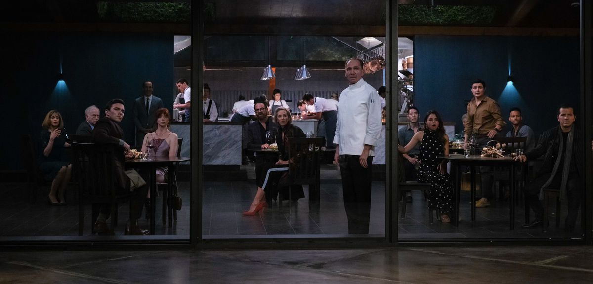 Lo chef Slowik si trova in una grande sala da pranzo con finestre, circondato da clienti del ristorante che sono tutti rivolti verso le finestre, guardando scioccato qualcosa fuori dallo schermo