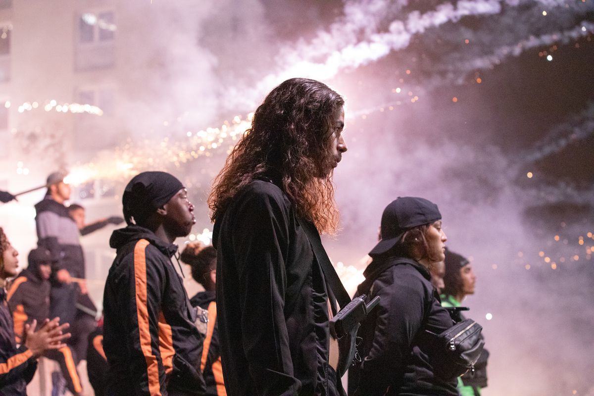Una foto di profilo laterale di un gruppo di giovani uomini in tuta nera con fumo e fuochi d'artificio in lontananza.
