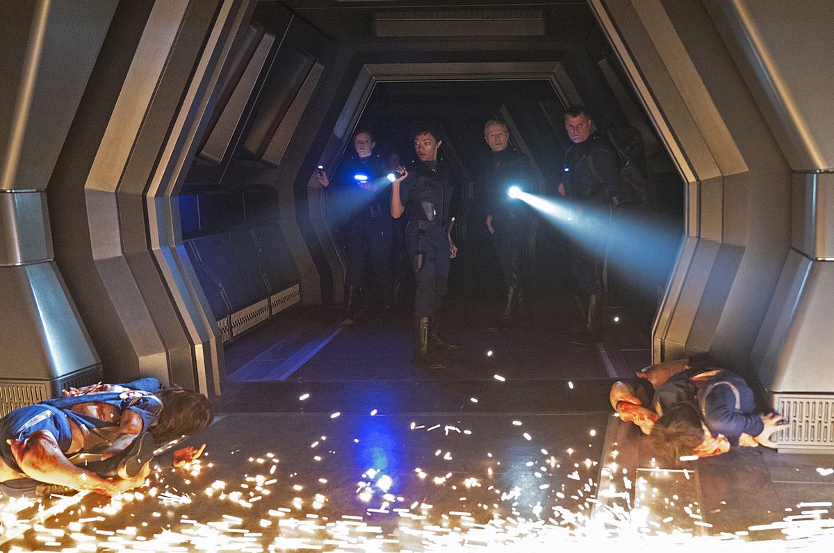 Quattro membri della Discovery in piedi in un corridoio con in mano torce elettriche e aggeggi, guardando corpi e scintille a terra di fronte a loro