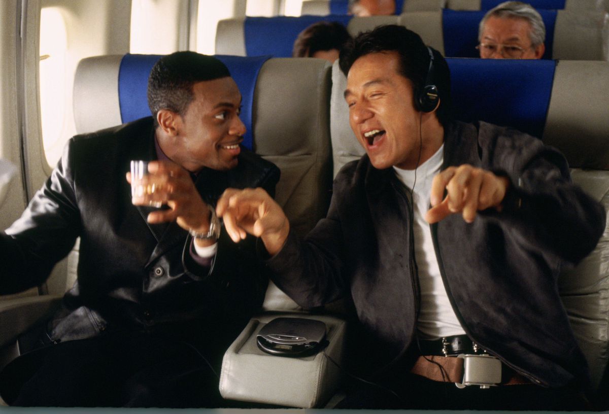 Due uomini seduti su un aereo commerciale, uno con in mano una tazza e l'altro con le cuffie e un aspetto visibilmente gioioso.