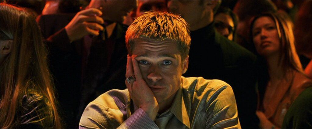 Un uomo (Brad Pitt) che guarda in avanti con la mano sulla guancia circondato dai partecipanti alla festa.