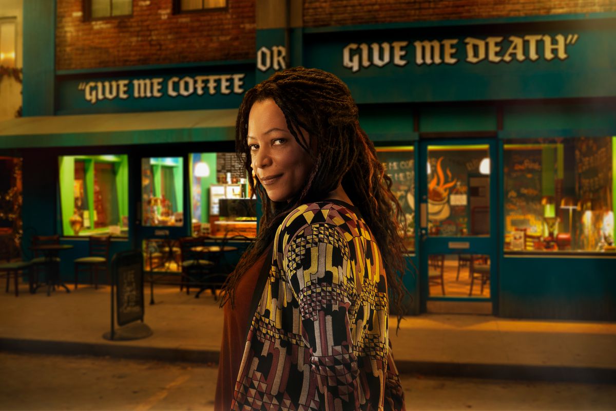 una donna con i dreadlocks scuri sorride compiaciuta davanti a un negozio che dice DAMMI IL CAFFÈ O DAMMI LA MORTE