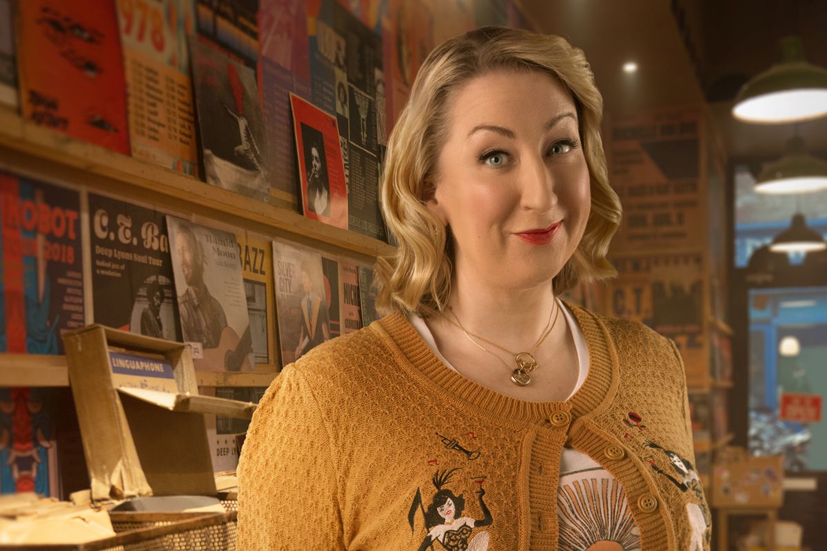 Maggie, una donna bionda con un cardigan giallo, in piedi in un negozio di dischi a Good Omens