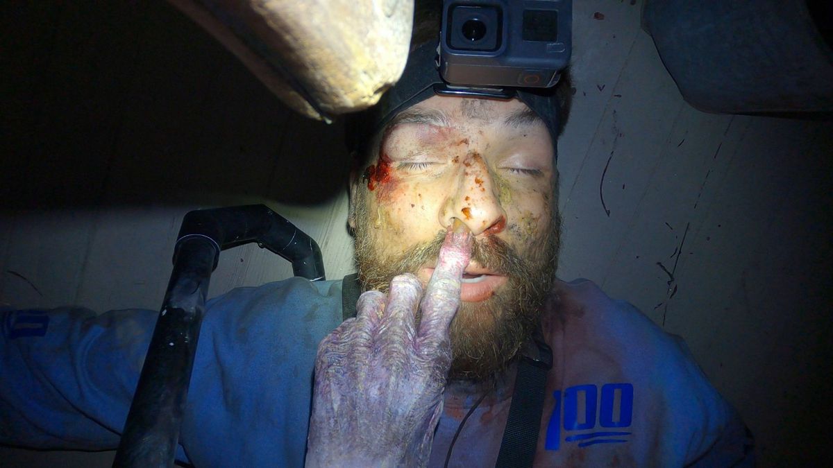 Un uomo che indossa una telecamera montata sulla testa giace privo di sensi su un pavimento di legno mentre una mano zombificata gli infila il dito in una delle narici.