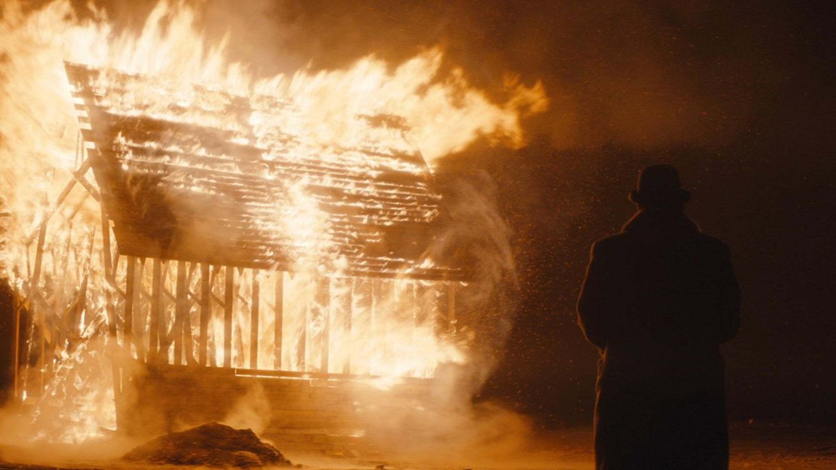 Una figura con un grande cappotto invernale e una bombetta si trova di fronte a un edificio incendiato nel cuore della notte.