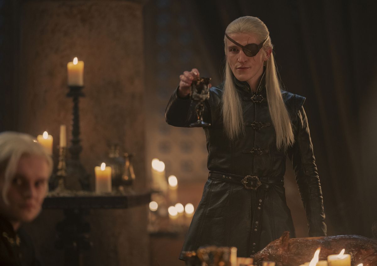 Ewan Mitchell nei panni di Aemond Targaryen in House of the Dragon alza un bicchiere in un brindisi beffardo con la benda sull'occhio