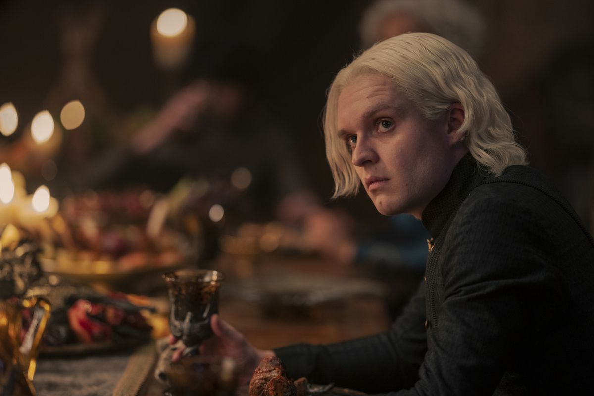Tom Glynn-Carry nei panni di Aegon II Targaryen seduto a un tavolo con un banchetto con in mano una coppa vincente in House of the Dragon