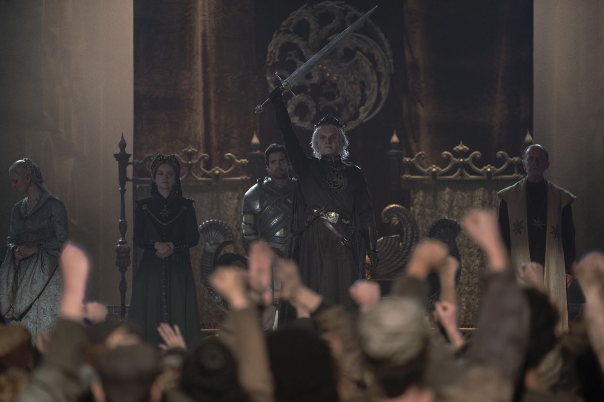 Re Aegon II (Tom Glynn-Carney) in piedi di fronte a una folla di persone esultanti di Westeros, tenendo la spada in aria trionfante 