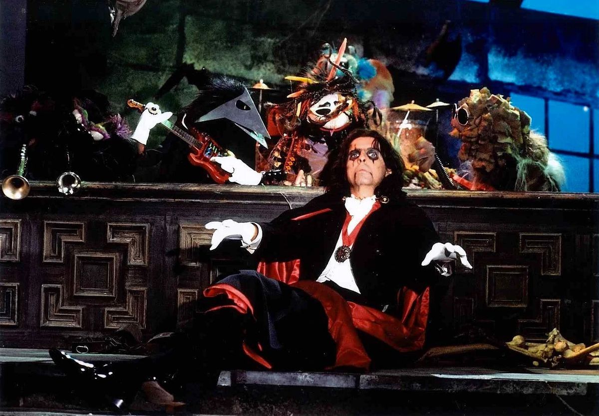 Alice Cooper, che indossa una matita per gli occhi sanguinante e vestita con un costume da vampiro e un mantello che ricorda il Dracula di Bela Lugosi, siede di fronte a una banda di burattinai.