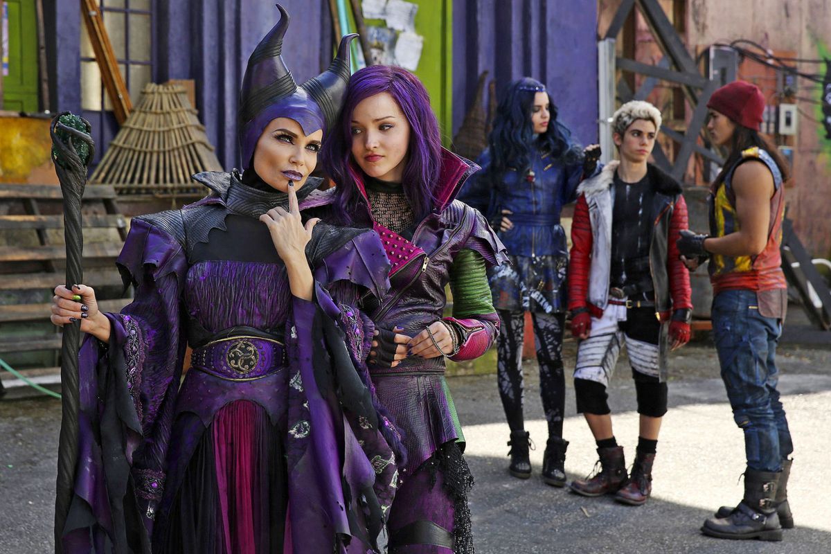 Una donna con un costume da strega viola con le corna è in piedi accanto a una ragazza con i capelli viola con una giacca da motociclista in pelle con altri tre bambini sullo sfondo che indossano costumi.