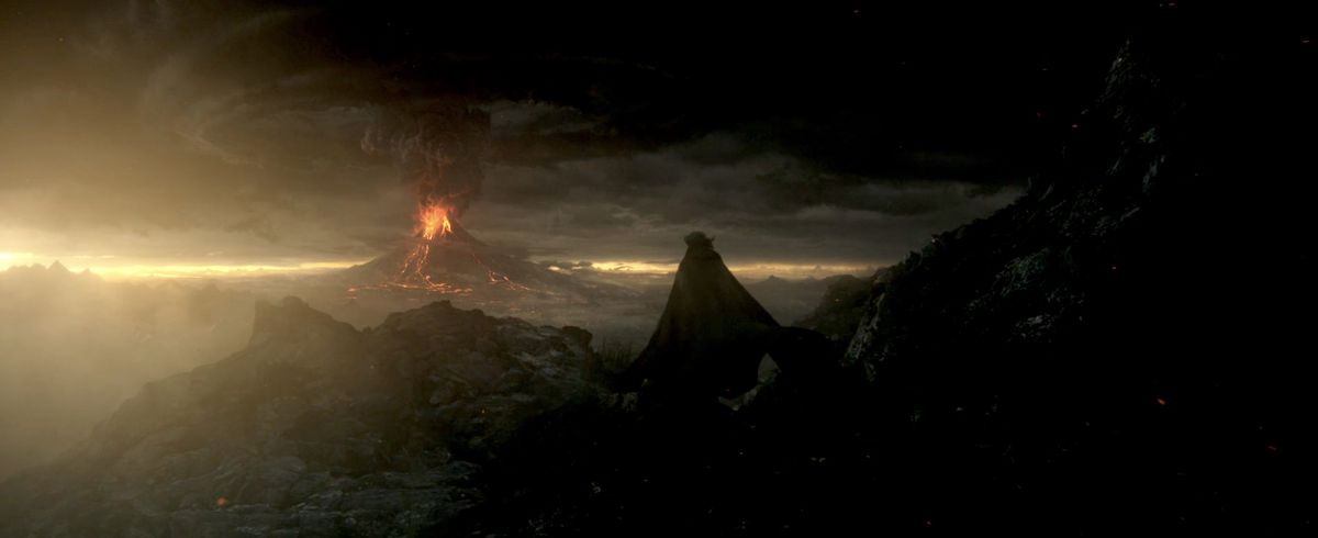 Sauron nel suo mantello nero sale su una scogliera per vedere il Monte Fato sparare della lava e del fumo nero che offusca il tramonto che sorge ne Gli anelli del potere
