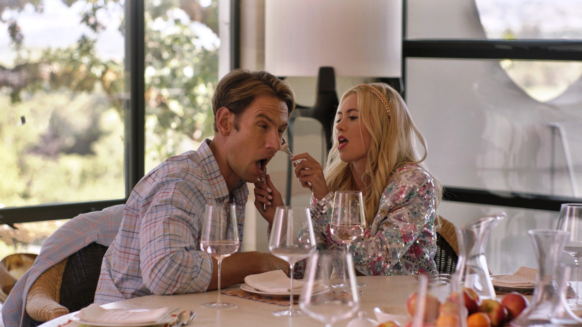 Una donna tiene una forchetta e un coltello all'orecchio di un uomo mentre fa una smorfia a un tavolo da pranzo circondato da bicchieri di vino.